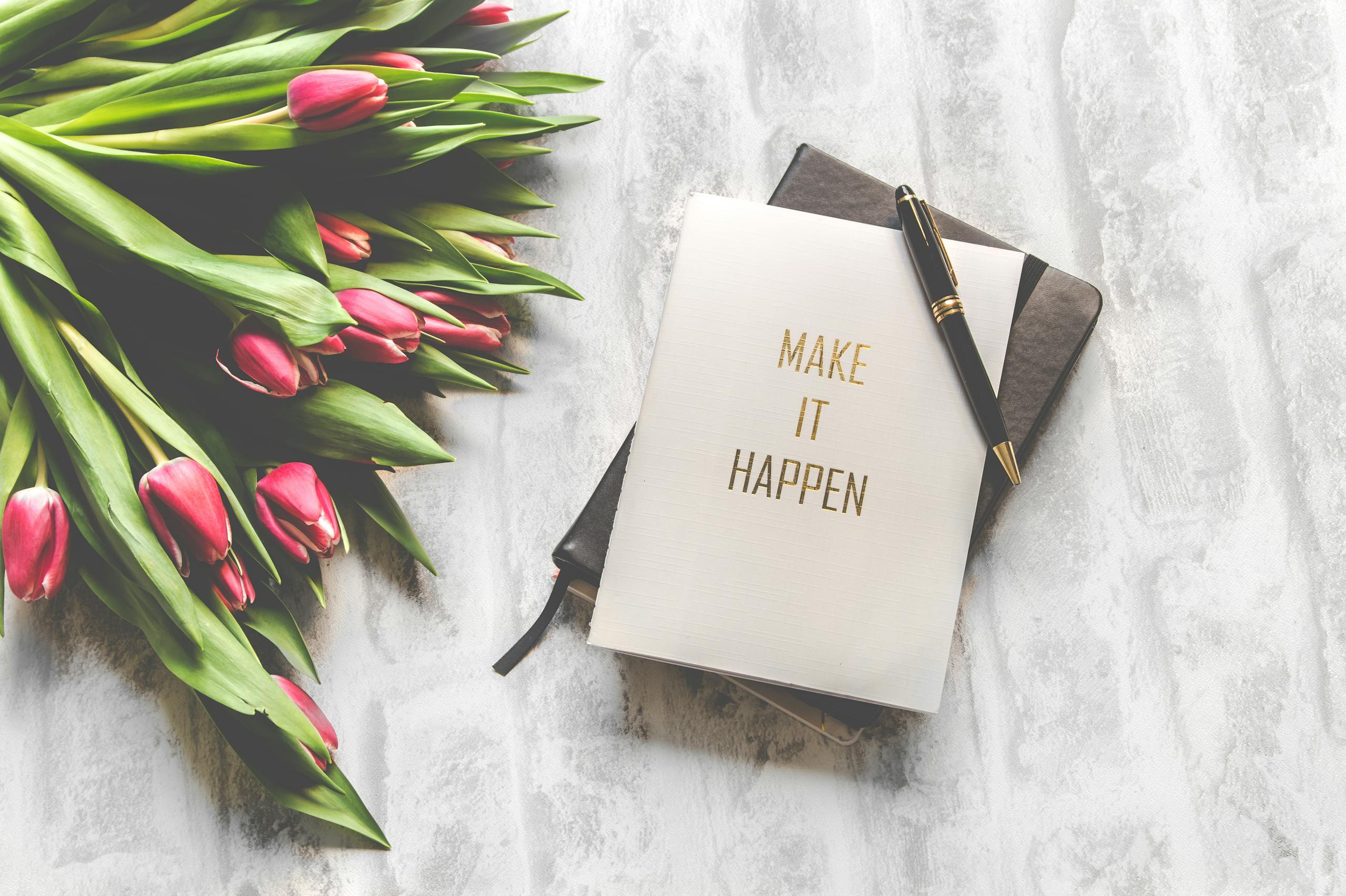 A notebook with 'Make It Happen' inscription beside a bouquet of pink tulips, capturing inspiration. Blog cover image for "How to Organise Your Life in a Weekend"